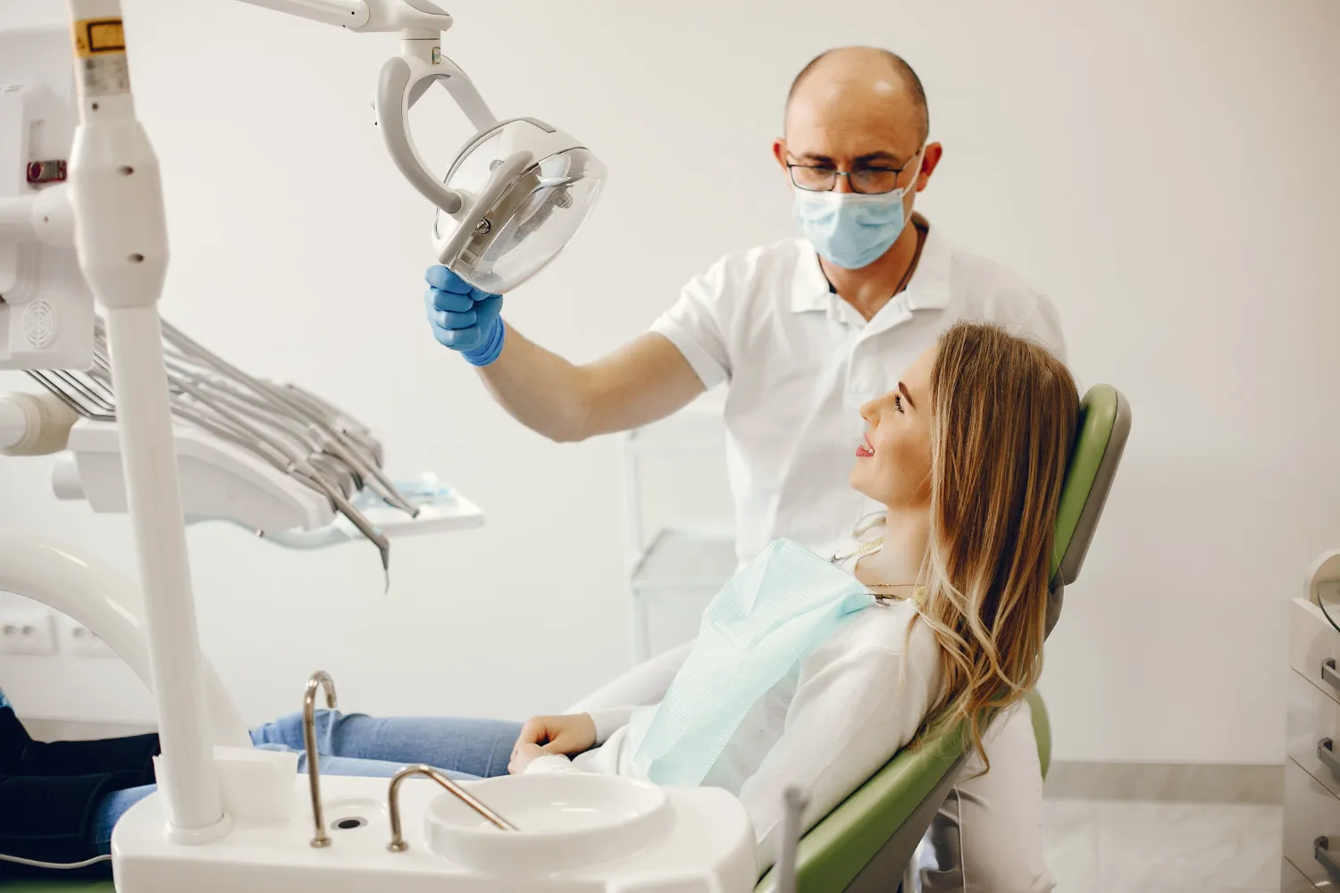 Ujęcie z bliska w jasnym gabinecie stomatologicznym. Mężczyzna dentysta, w maseczce chirurgicznej, okularach i niebieskich rękawiczkach, koryguje położenie dużej lampy zabiegowej nad głową leżącej na fotelu kobiety. W tle widoczne są sterylne urządzenia stomatologiczne i szafka.